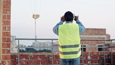 Rear-of-the-African-American-man-architect-or-foreman-in-VR-glasses-standing-on-the-roof-at-the-building-site-and-having-headset-as-watching-virtual-tour-of-future-architecture.-Outside.-New-modern-technologies.-Back-view.